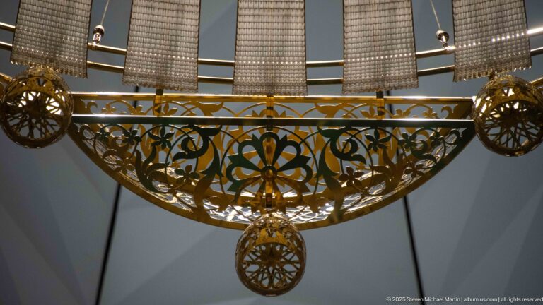 Gilded chandelier detail with pierced floral scrollwork, circular ornaments, and a hanging finial.