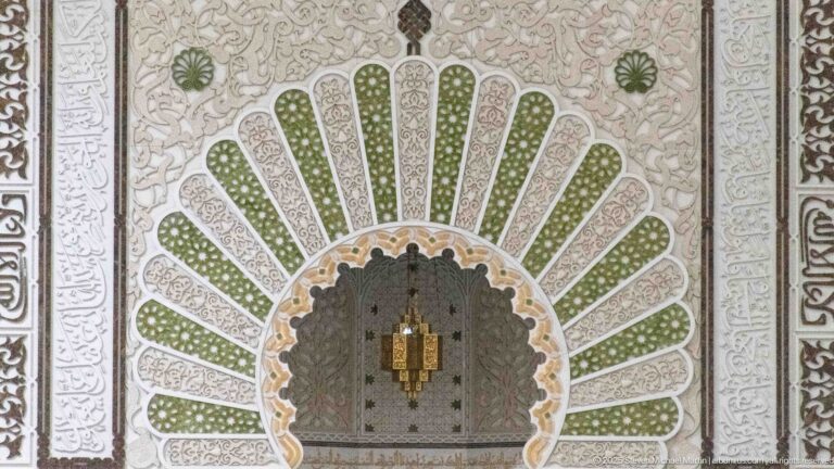 Mihrab detail with green radiating panels, pale arabesque relief, dark borders, and a small hanging lamp.