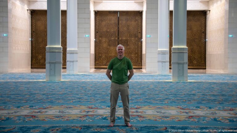 Man in a green shirt stands on the blue carpet between white columns and large bronze-toned doors.