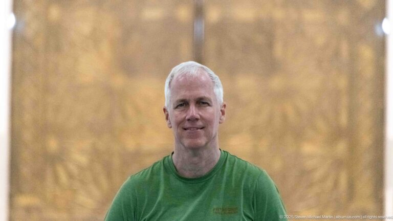 Close portrait of a gray-haired man in a green shirt against a softly blurred golden interior.