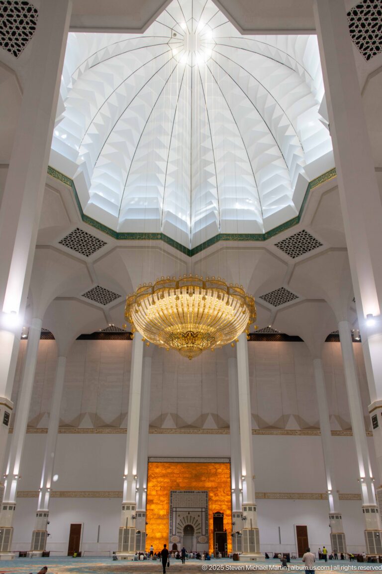 Large chandelier and faceted ceiling recess align above the distant glowing mihrab wall between tall white columns.