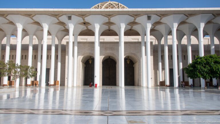 White arcade fronts pale stone walls and tall arched doors across a reflective mosque courtyard.