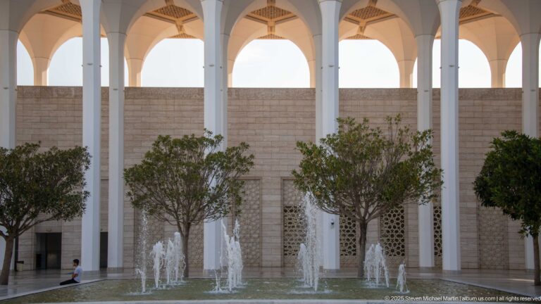 Water jets rise from a courtyard fountain between trees and tall white mosque arcades.