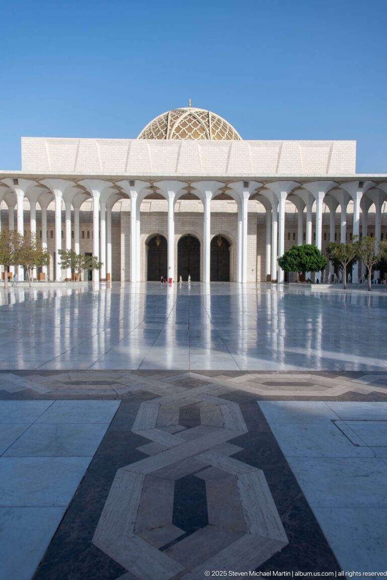 Geometric paving leads across a broad courtyard toward a white arcade and patterned dome.