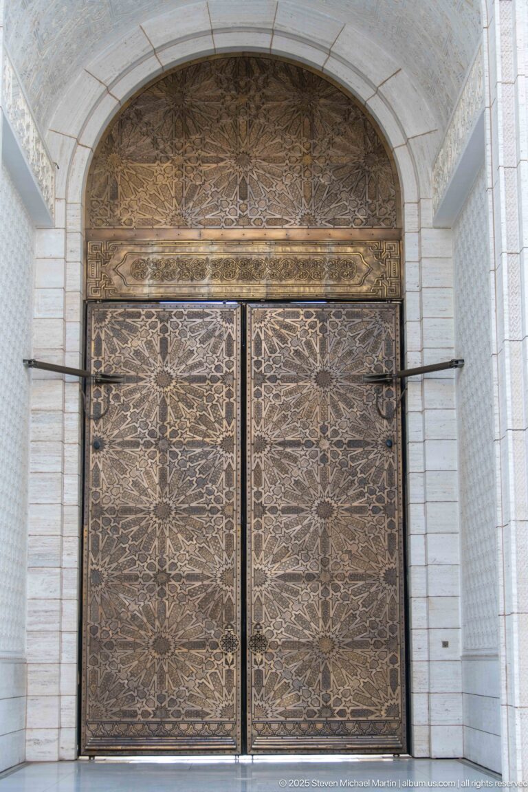 Large ornate metal double door with dense symmetrical patterns beneath a decorated arched panel.