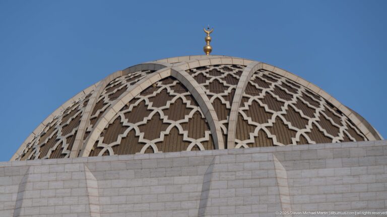 Ribbed patterned mosque dome with geometric lattice panels and a gilded crescent finial against blue sky.