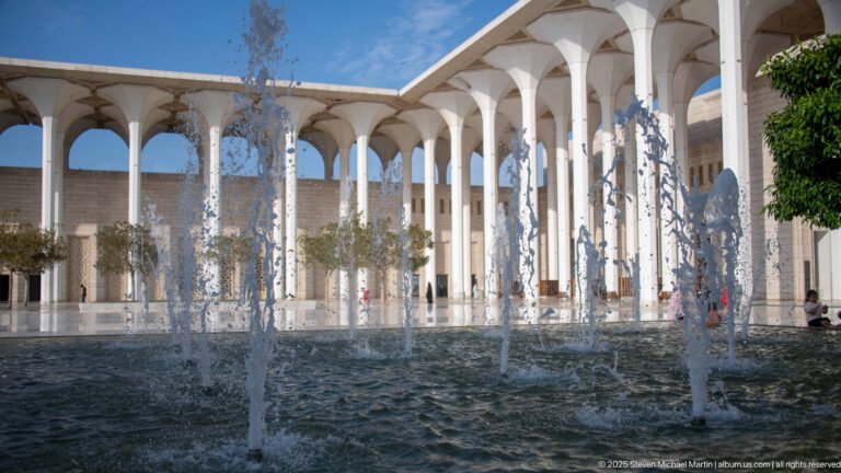 Fountain jets rise in the foreground with white arcades and courtyard trees behind.
