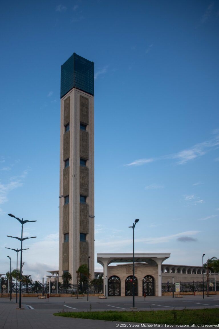 Tall rectangular minaret with recessed panels and a dark cap rises beside a low mosque entrance.