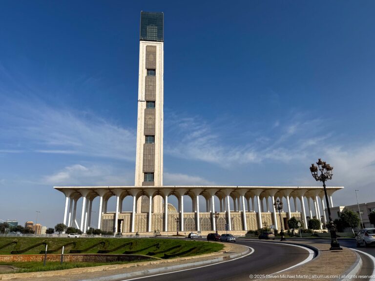 Very tall rectangular minaret rises above a long white arcade beside a curved roadway.