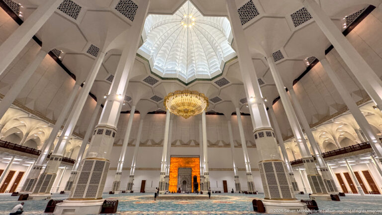 Wide interior with tall white columns converging toward a chandelier, faceted ceiling, and glowing mihrab wall.