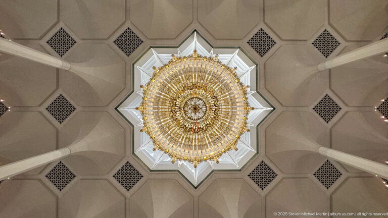 Large circular gilded chandelier centered within a bright star-shaped ceiling recess surrounded by pale geometric panels.