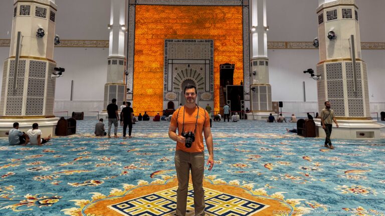 Man in an orange shirt with a camera stands on the mosque carpet before the glowing mihrab wall and seated worshippers.