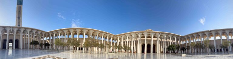 Ultra-wide mosque courtyard with curving white arcades, polished paving, trees, minaret at left, and dome near center.