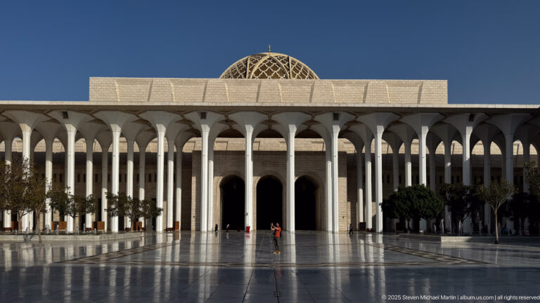 Patterned dome above a long white arcade and three tall dark entrances across the reflective mosque courtyard.