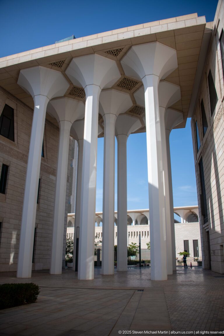 Broad paved forecourt beneath four white flared columns connecting stone-clad wings of the mosque.
