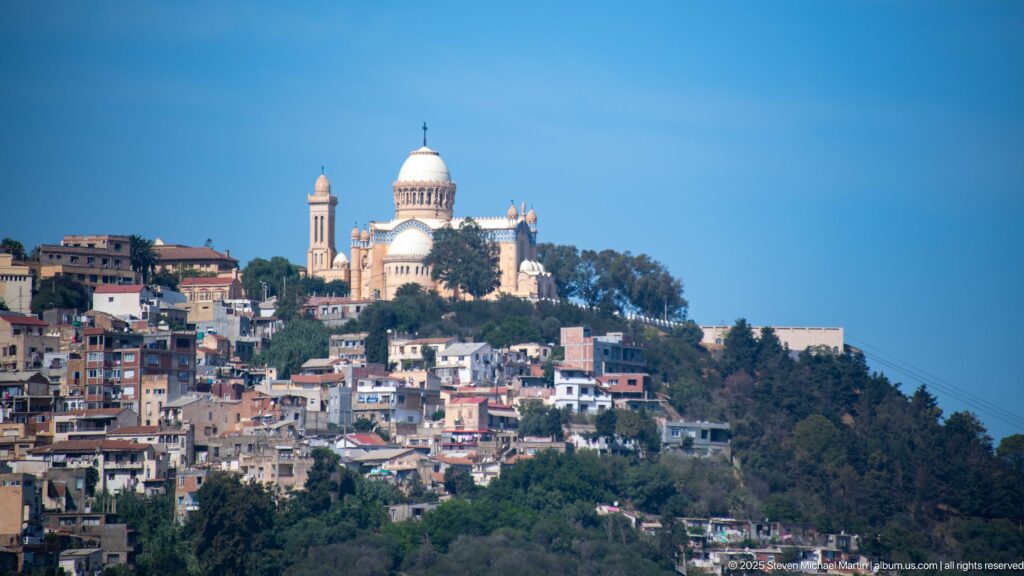 Notre-Dame d’Afrique basilica above hillside neighborhoods in Algiers, with its dome and towers prominent against a clear blue sky.