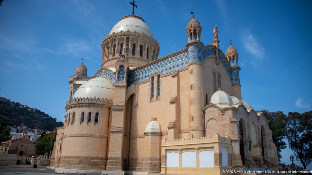 Close exterior view of Notre-Dame d’Afrique with rounded apses, domes, corner turrets, and blue tile bands.