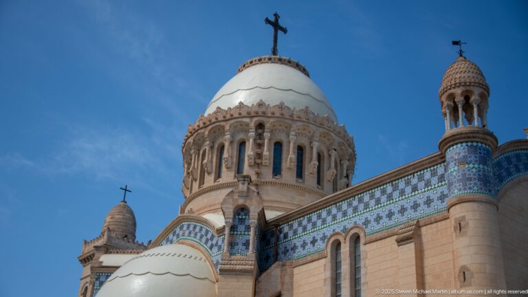 Upper exterior of Notre-Dame d’Afrique showing the main dome drum, arched openings, blue tile bands, and a smaller turret.