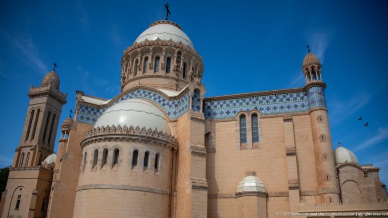 Frontal exterior of Notre-Dame d’Afrique with rounded apse, central dome, blue tile bands, bell tower, and small turrets.