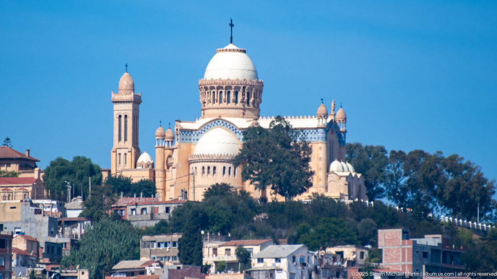 Closer view of Notre-Dame d’Afrique showing the central dome, bell tower, small domed turrets, and blue decorative tile bands.