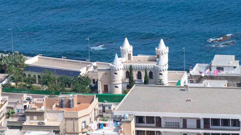 White coastal building with four corner towers near the Mediterranean shoreline below Notre-Dame d’Afrique.