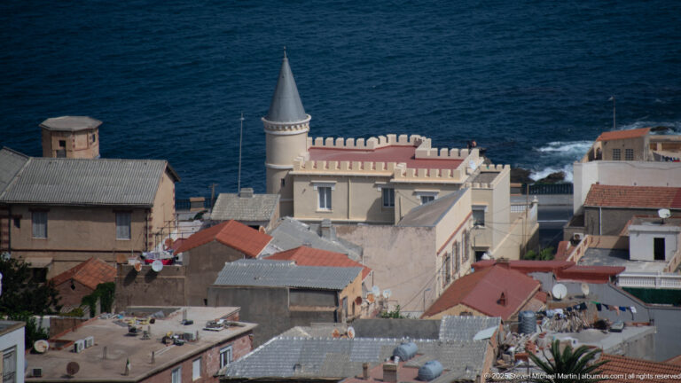 Turreted pale residence with crenellated parapet among Algiers rooftops beside the Mediterranean.