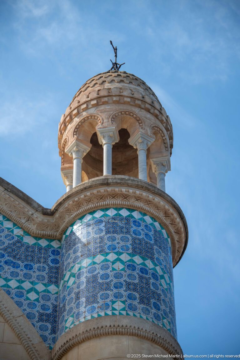 Small open turret with columns and cross above blue patterned tilework on Notre-Dame d’Afrique.