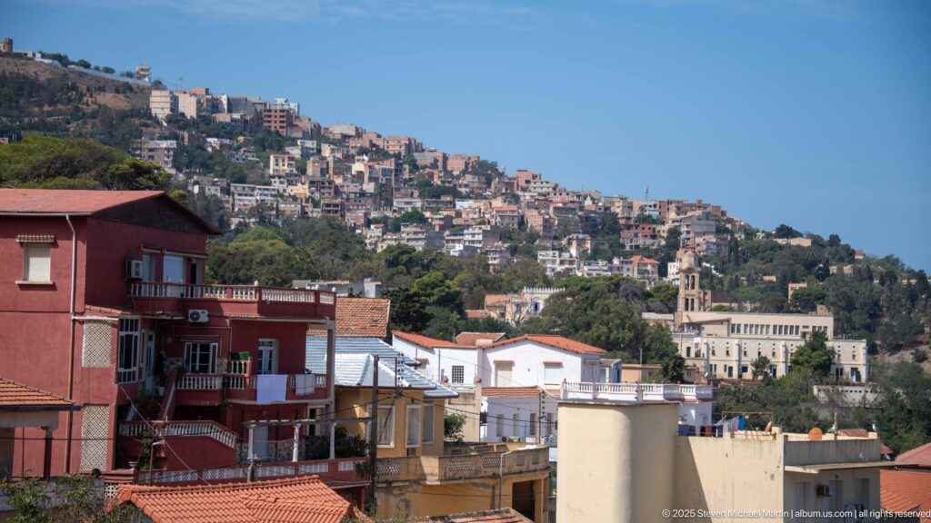 Layered Algiers cityscape with lower rooftops and dense hillside neighborhoods climbing the slope near Notre-Dame d’Afrique.