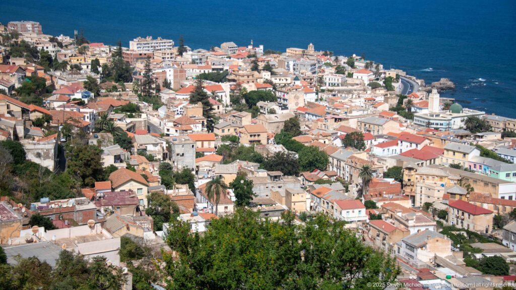 Elevated view over a dense coastal quarter of Algiers with red-roofed homes descending toward the Mediterranean.