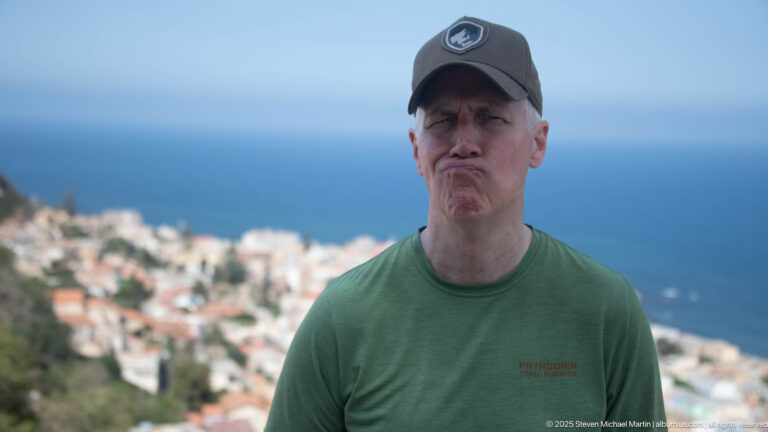 Man in a dark cap and green T-shirt posing at a hilltop viewpoint above coastal Algiers and the Mediterranean.
