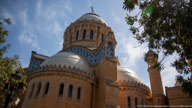 Notre-Dame d’Afrique seen from below through trees, with rounded apses, blue tile bands, turrets, and central dome.