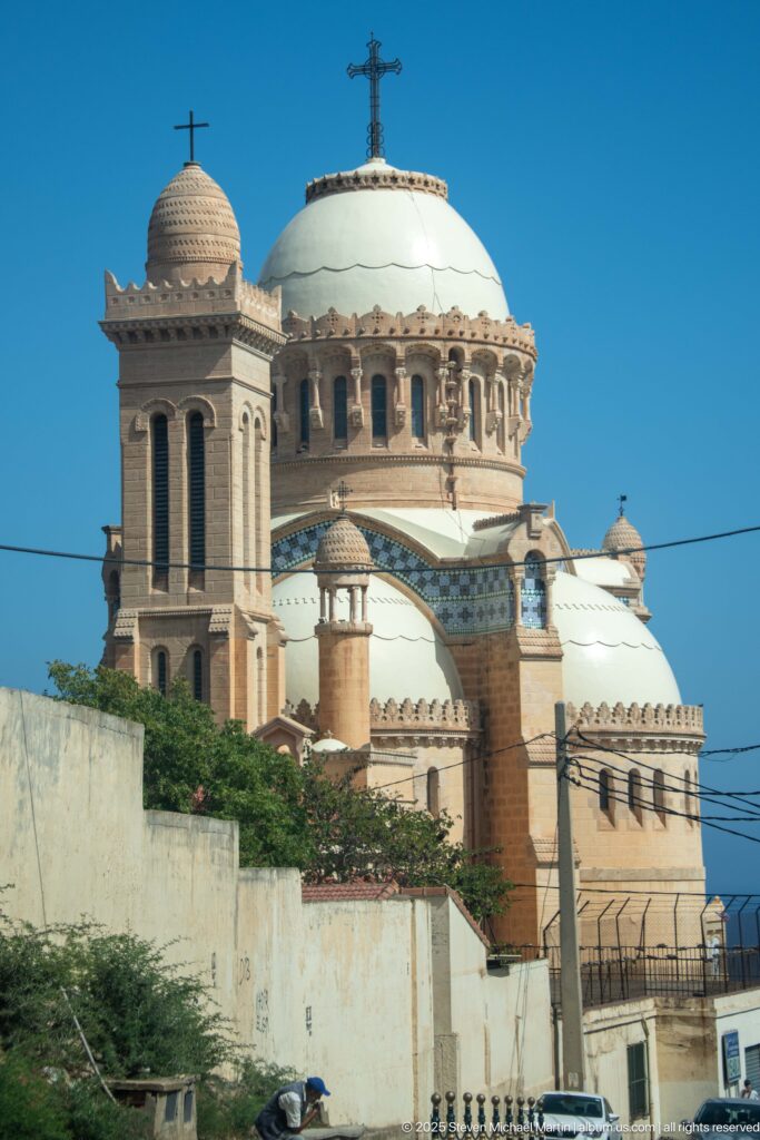 Side view of Notre-Dame d’Afrique with layered domes, a corner tower, pale stone walls, and overhead utility lines.
