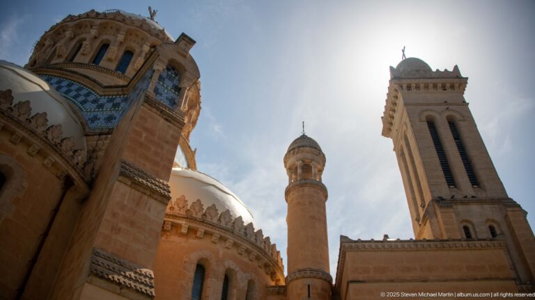 Backlit low-angle view of Notre-Dame d’Afrique with square bell tower, domed roofs, slender turret, and blue tile bands.