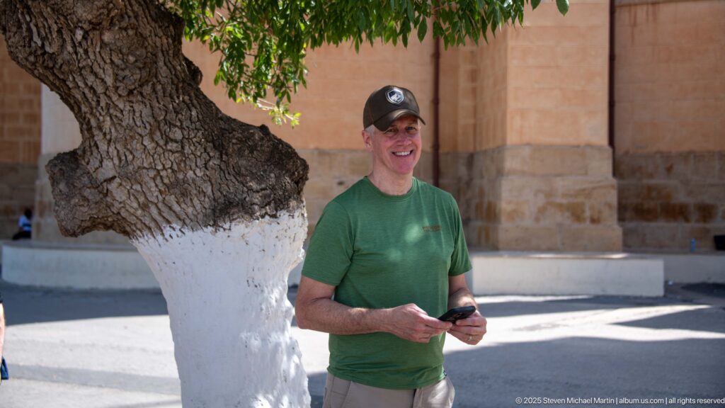 Smiling man in a green T-shirt and dark cap standing beside a whitewashed tree near Notre-Dame d’Afrique.