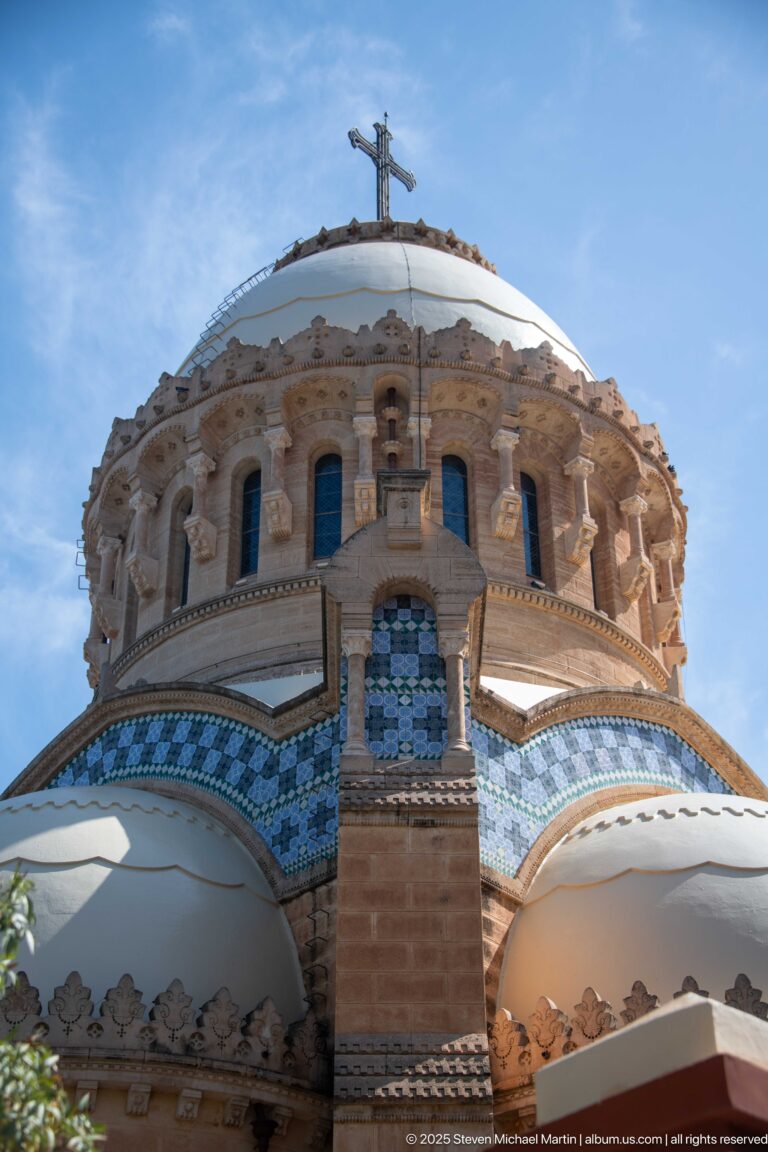 Close low-angle view of Notre-Dame d’Afrique’s central dome drum, arched openings, cross, blue tile bands, and rounded roofs.
