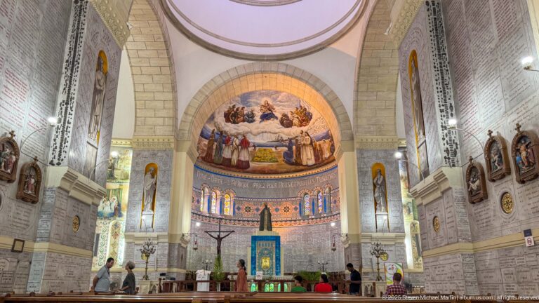 Centered interior of Notre-Dame d’Afrique with pews, altar, apse mural, stained glass, mosaics, and visitors near the sanctuary.