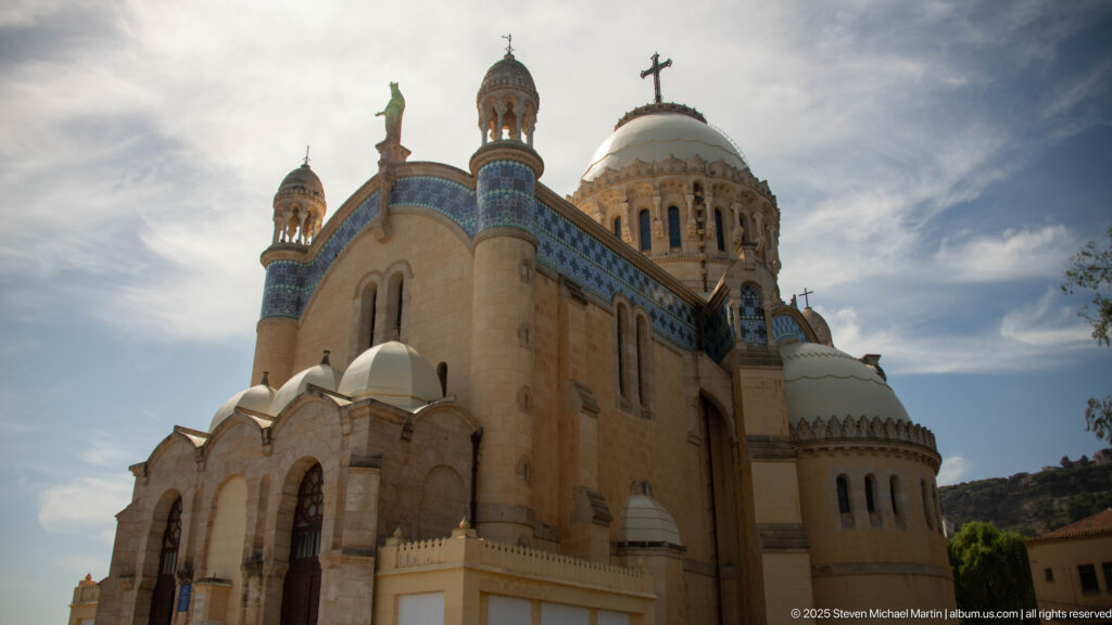 Low-angle view of Notre-Dame d’Afrique showing rounded apses, arched windows, tiled bands, turrets, and the main dome.