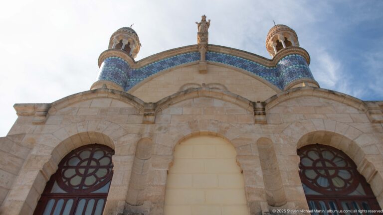 Exterior detail of Notre-Dame d’Afrique with three stone arches, circular window grilles, blue tilework, small turrets, and a roofline statue.