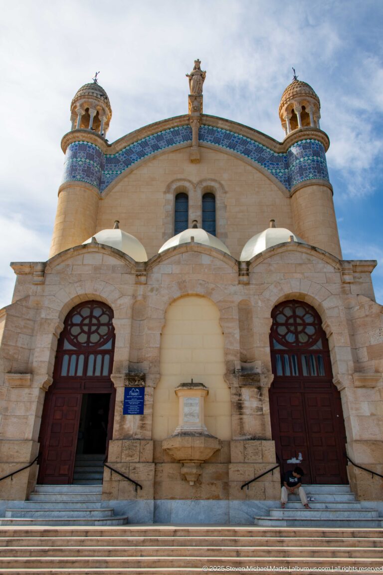 Front façade of Notre-Dame d’Afrique with steps, paired entrances, rounded arches, circular glazing, tilework, and rooftop sculpture.