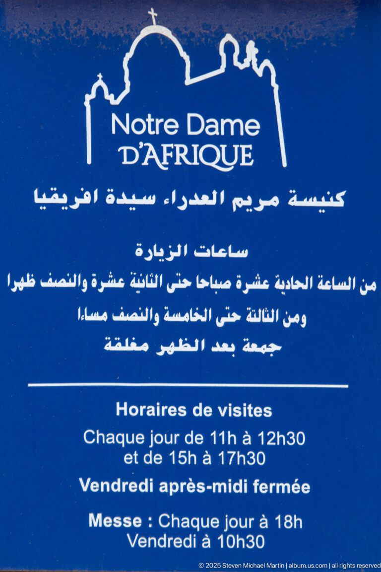 Blue Notre Dame d’Afrique visitor sign with French and Arabic text, basilica logo, visiting hours, and Mass times.