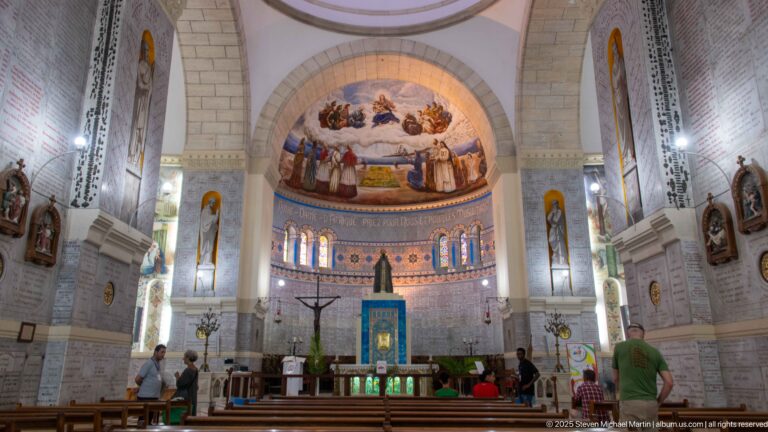 Interior of Notre-Dame d’Afrique with pews, broad arches, altar, mosaic apse, stained glass, and visitors near the sanctuary.