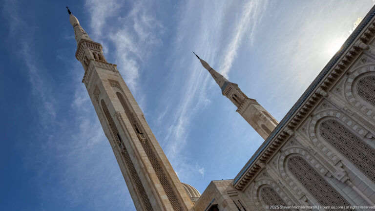 Low-angle view looking up at the mosque’s twin minarets beside an ornate white façade under blue sky.
