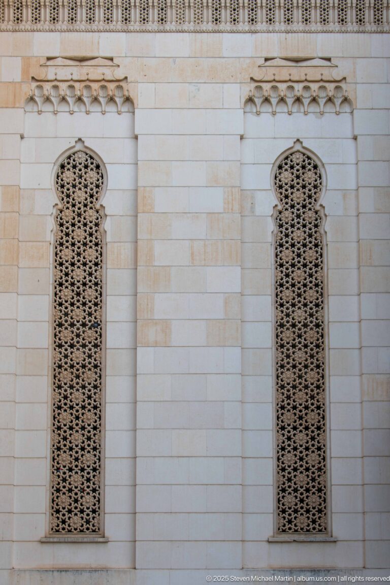 Two tall arched windows with dense geometric screens set symmetrically in the mosque’s pale stone wall.