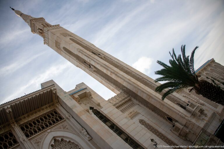 Low-angle view of a mosque minaret rising above ornate façade details and a palm tree.