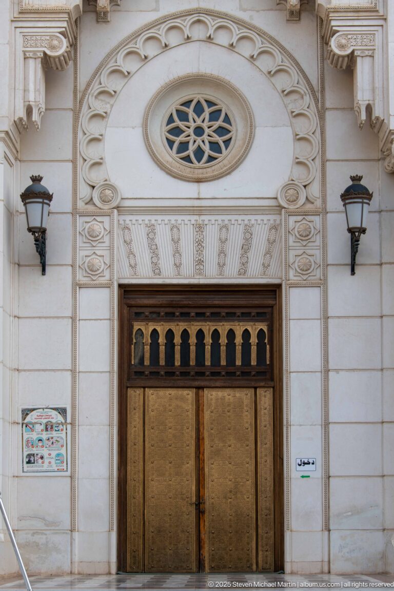 Bronze-toned double door beneath a circular rosette and carved arch bands, flanked by wall lamps.