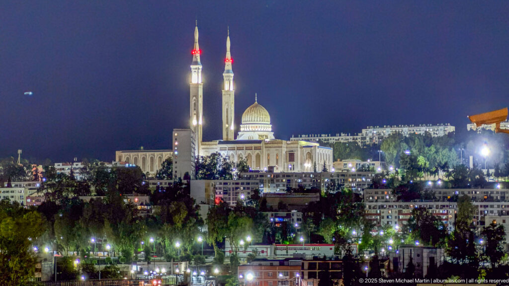 Illuminated Amir Abdel Kader Mosque with twin minarets and central dome rising above Constantine at night.