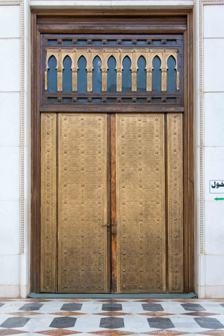 Close view of patterned bronze-toned double doors beneath a dark wooden transom with lobed openings.