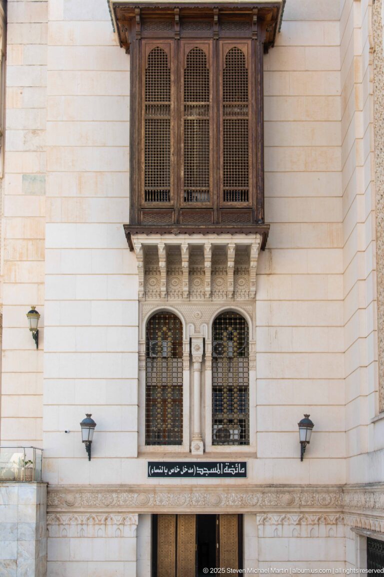 Projecting wooden window above paired arched windows, an Arabic sign, and a secondary doorway on the mosque façade.