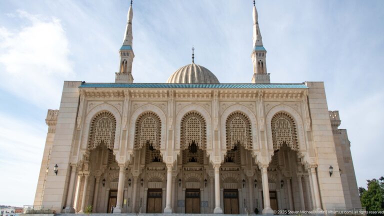 Frontal mosque façade with broad steps, five arched entrance bays, central ribbed dome, and twin minarets.