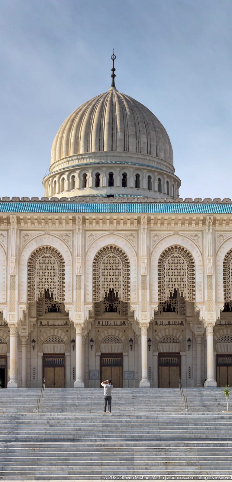Large ribbed mosque dome above an ornate entrance arcade and broad steps, with one person crossing the foreground.
